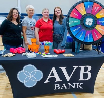 Four women standing around a table with a prize wheel and promo items for a hiring event. 